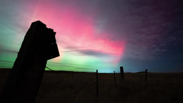 Green and red northern lights dancing behind a weathered fence post and barbed wire fence on the plains, Montana, time lapse