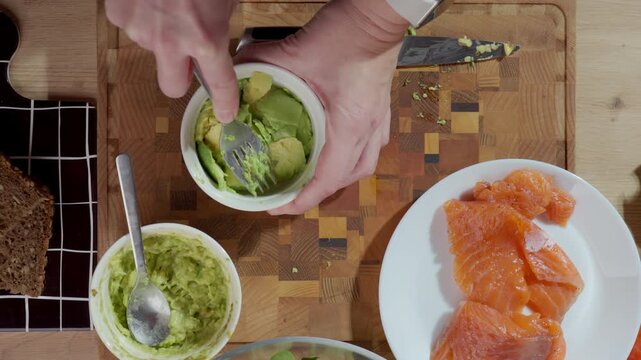 Hands mash avocado in bowl on cutting board beside salmon and bread. Concept of healthy breakfast preparation and homemade recipe process.