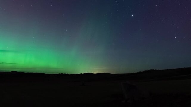 Green northern lights glowing on the horizon as the moon rises over dark rolling hills, Montana, time lapse