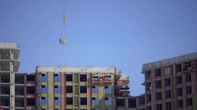 Large tower crane lifting a heavy load of building materials against a clear blue sky, moving it towards an unfinished multi-story apartment block with external wall insulation