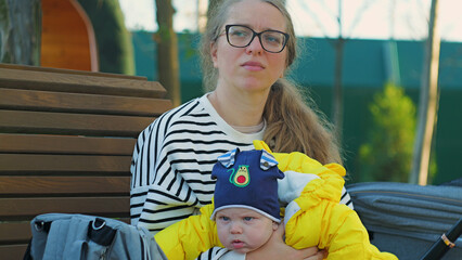 Candid outdoor portrait of a young mother holding her baby boy. She has a pensive look while...