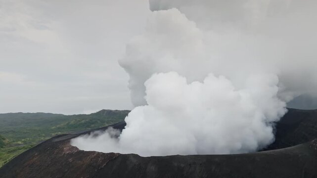 Yasur Mountain volcano on Tanna Island, Vanuatu, actively erupting, sending a thick column of white ash and smoke into the gray sky, with green vegetation visible in the background. Aerial panorama