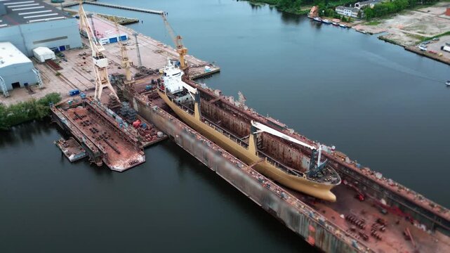 Aerial drone shot circling a massive dry dock. Large vessel is propped up inside, out of the water, undergoing heavy maintenance or repairs. Shipping, logistics, naval engineering, or construction. 
