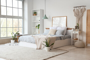 Interior of light bedroom with cozy bed, bedside table, houseplants and shelving unit