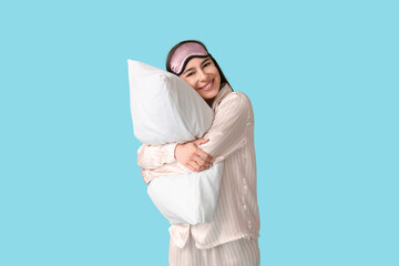 Young woman in pajamas with sleeping mask hugging pillow on blue background