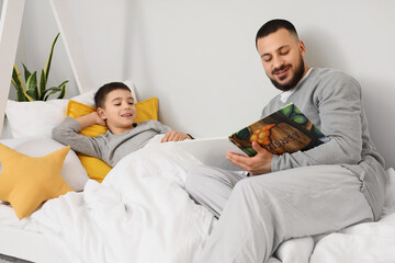 Naklejka premium Happy father in pajamas reading book for his son on bed at home