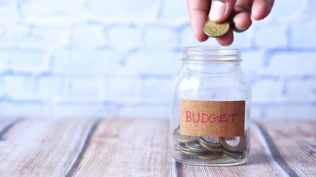 Person hand putting golden coins into glass jar with budget label on wooden table against brick wall background