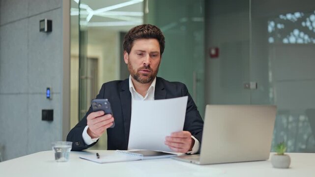 Businessman analyzes important data. Information cross-referencing on smartphone and document in modern office. Expresses seriousness, focus. Task requires concentration.