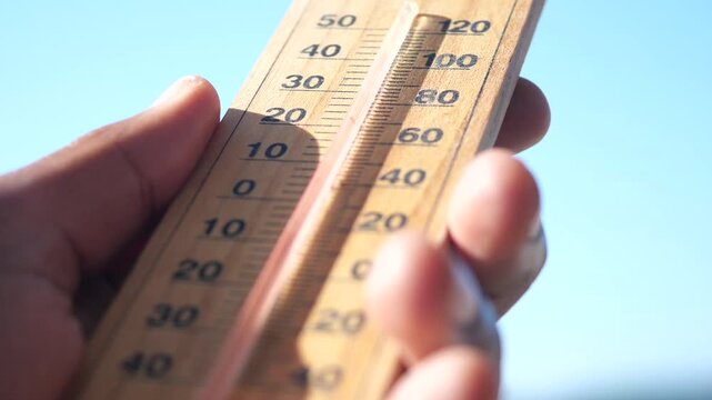 A close up view of a hand holding a wooden thermometer against a clear blue sky, showing the temperature rising due to the hot weather