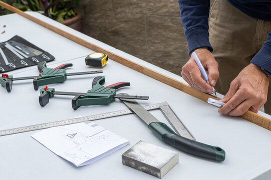 Handyman hands holding a pencil and marking a wooden plank, working on a diy project on a white workbench with various tools like clamps, measuring tape, and a design plan