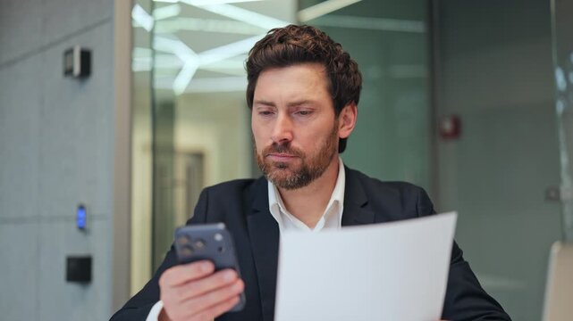 Professional deeply focused, engaged with phone and documents. Displays intense concentration and serious dedication. Captures efficient workflow, reviewing important business information calmly.