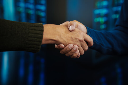 Close-up of two businesspeople shaking hands in a data center or server room with blue lights and blurred racks, symbolizing partnership, agreement, cybersecurity and technology collaboration.