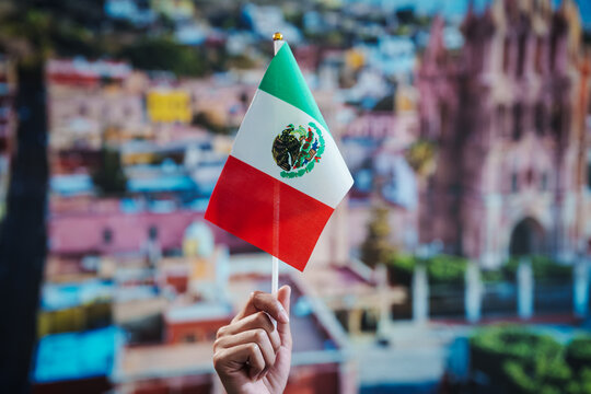 Hand holding small mexico flag with cityscape and cathedral blurred in background, patriotic travel concept, shallow depth of field.