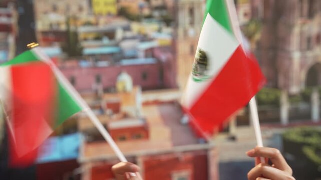 Hands waving small mexican flags against blurred colorful cityscape background, festive patriotic celebration with shallow depth of field.