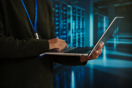 It professional using laptop in data center server room, cybersecurity and network admin concept with blue lights and racks in background