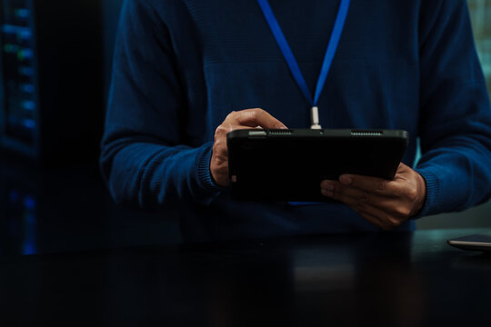 Close-up of IT technician with lanyard using a tablet in a server room, blue lighting, data center racks blurred in background; cybersecurity and system monitoring concept.