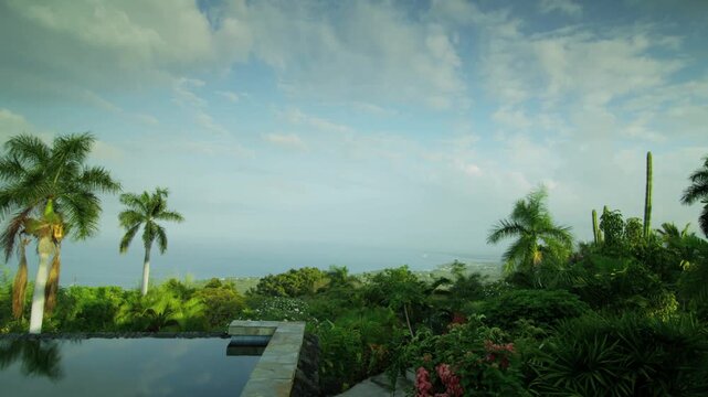 Time-lapse footage of a tropical landscape featuring a reflective infinity pool in the foreground, lush greenery, and palm trees with the ocean visible in the distance. The partly cloudy sky.