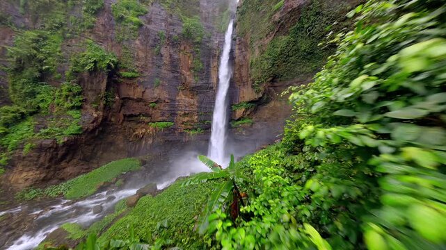 Cienmatic FPV drone flight through lush tropical jungle toward Kapas Biru waterfall in East Java, Indonesia. Dramatic rainforest landscape with tall cascade and mist in Southeast Asia.