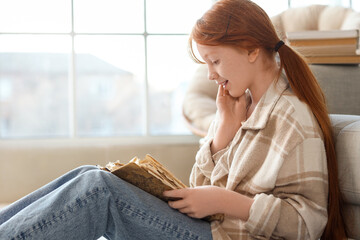 Naklejka premium Cute teenage happy redhead girl with adventure book sitting on floor near window at home