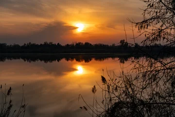 A serene and beautiful sunset over a tranquil lake, featuring the sun in a distinct heart shape amidst warm, glowing clouds. The still water perfectly mirrors the vibrant sky and the unique sun. © Dina Luk