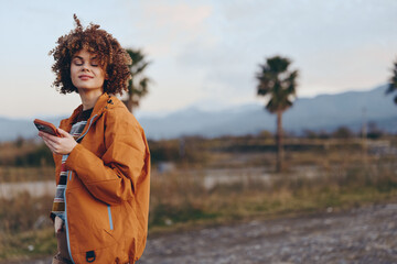 Smiling woman with curly hair wearing rainbow sweater and orange jacket holding smartphone outdoors...