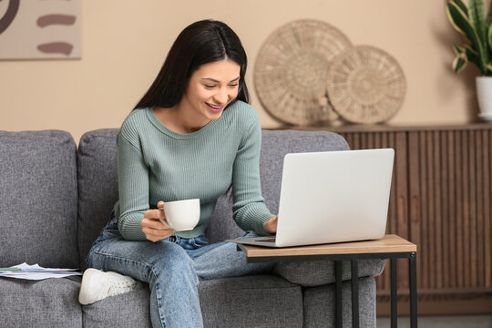 Beautiful young woman with cup of coffee and laptop sitting on sofa in living room