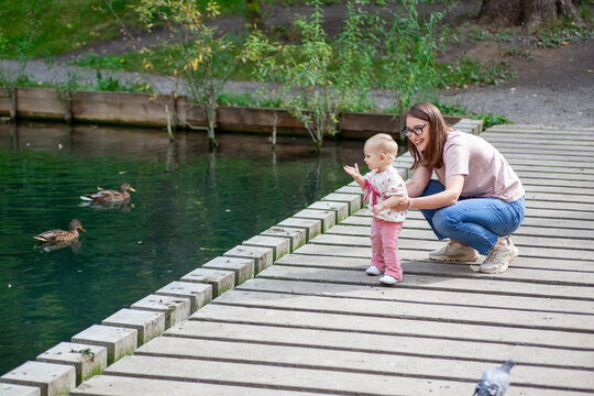 A mother and her young child are standing by a pond, watching ducks swim. The child is pointing at the ducks with excitement while the mother smiles and encourages.