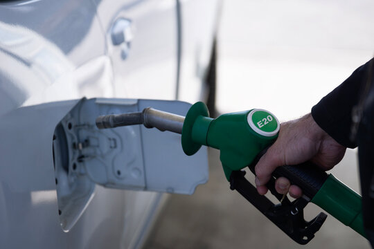 man refilling car with e20 biofuel at gas station, hand holding green fuel nozzle with e20 label close up, white vehicle parked outdoors, concept of oil industry, energy sector, fuel economy