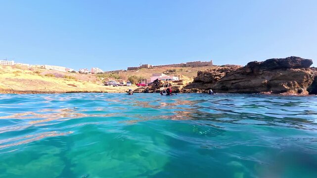Turquoise Mediterranean Waters Glistening Under Summer Sun with Scuba Divers Emerging Near Golden Rocky Coastline and Ancient Fortification in Background Captured from Low Water Surface Camera Angle
