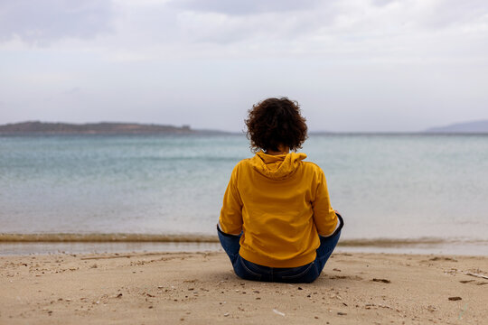Back view of woman in yellow hoodie sitting in lotus pose on beach