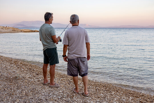 Two men fishing and talking on rocky beach rear view