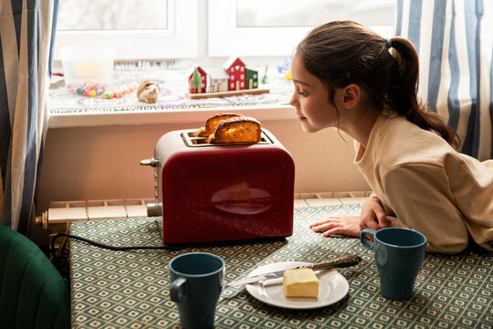 Girl looking at toast popping out of red toaster on kitchen table.