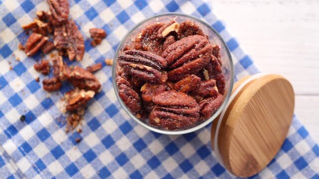 Top view of candied pecan nuts in glass jar on checkered napkin over white wooden background