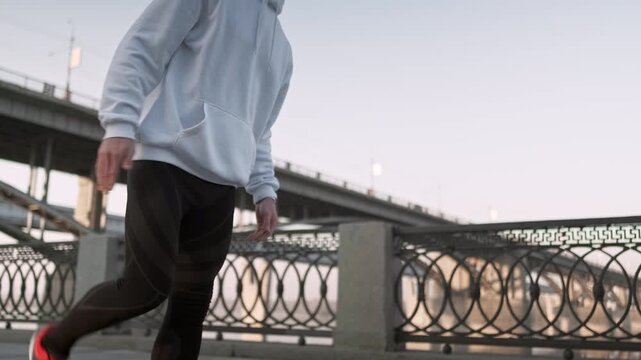 Young athlete wearing a white hoodie and black leggings stretches his legs near a bridge, preparing for a morning run. The calm riverside environment emphasizes outdoor fitness, healthy lifestyle, and