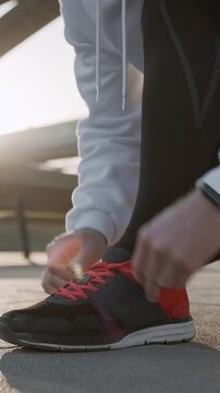 Sportsman in athletic clothing fastening his running shoes beneath a bridge at sunrise. The image captures preparation for a morning run, emphasizing urban fitness, motivation, and active lifestyle.