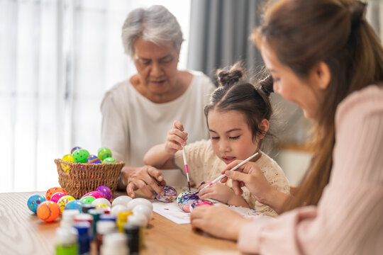 Asian little girl painting colorful easter eggs with brush while grandmother helping her at kitchen table, Three generations family spring holiday celebration, creative art craft learning, easter day
