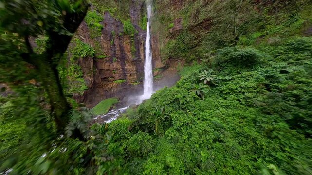 Cienmatic FPV drone flight through lush tropical jungle toward Kapas Biru waterfall in East Java, Indonesia. Dramatic rainforest landscape with tall cascade and mist in Southeast Asia.
