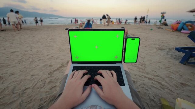POV working on laptop with smartphone green screen on beach