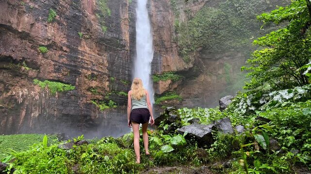Woman standing in front of Kapas Biru waterfall surrounded by lush tropical jungle in East Java, Indonesia. Scenic rainforest landscape with dramatic cascade in Southeast Asia.