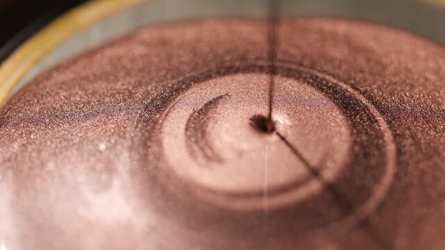 Continuous stream of dark golden paint pouring into viscous pool creating concentric circular ripples and showcasing specialized sandy texture