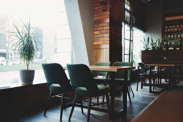 Fototapeta premium Inside a cafe, a table with four chairs sits next to a large window. Sunlight streams in. Plants decorate the space. More tables and chairs are visible in the background.
