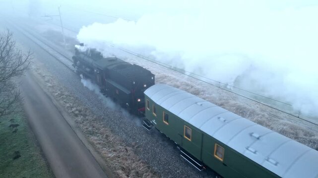 A vintage steam train, in retro style, moves through a misty landscape, emitting thick white smoke. The old-fashioned rail transportation evokes nostalgia, with green passenger cars trailing behind.