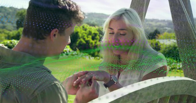 Man holding woman's left hand placing ring proposing on swing, green purple dots accenting moment