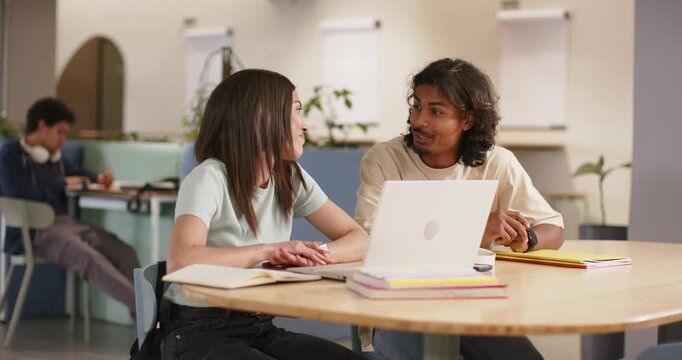 Two students conversing, shifting from pen and yellow notebook to typing on laptop at study lounge