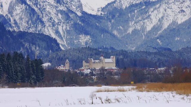 Cinematic winter video with a smooth horizontal panning shot across a snow-covered valley, revealing Hohenschwangau Castle and the village beneath the majestic Bavarian Alps, Germany.