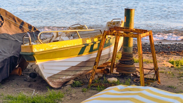 A yellow and white boat sits on a sandy beach near the ocean at sunset A metal capstan structure and ropes are nearby in this coastal scene.