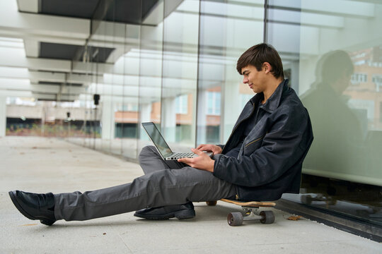 Young man relaxing on skateboard using laptop by glass building