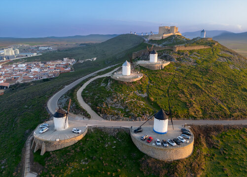 Aerial view of iconic windmills standing proudly on a hilltop, their white bodies contrasting with the dark caps and the winding road, Consuegra, Castile-La Mancha, Spain.