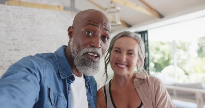 Diverse couple tapping smartphone, recording selfie while leaning in bright living room, smiling