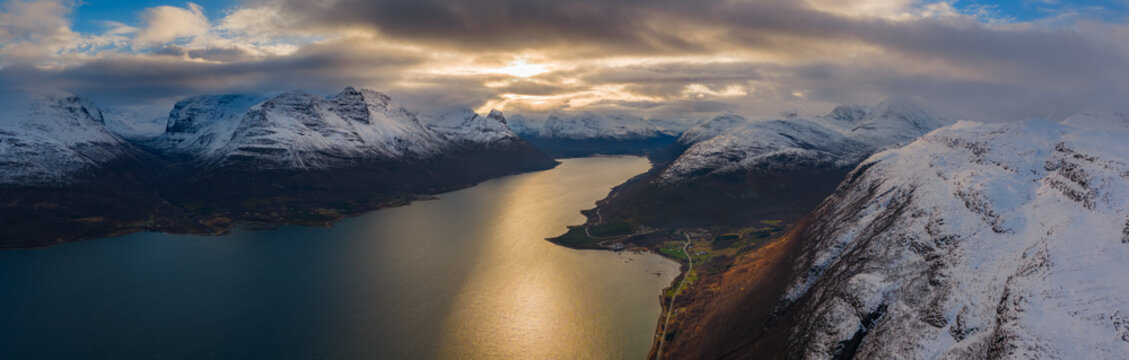 Aerial view of golden sunlight piercing through the clouds, reflecting off the still, dark waters amidst snow-capped mountains, Skibotn, Troms, Norway.